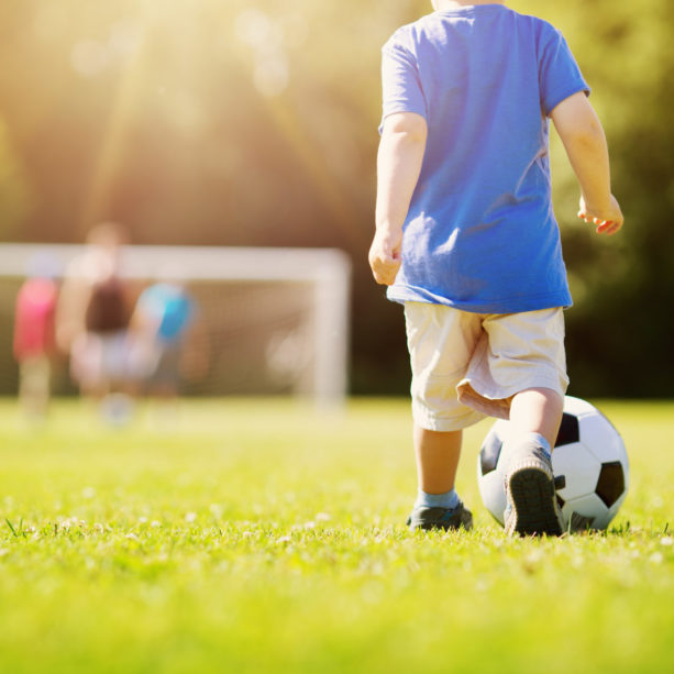 Little boy playting football on the field with gates. Active child in summer with ball
